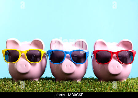 Drei rosafarbenen Spardosen mit Sonnenbrille auf Gras mit blauem Himmel. Studio shot mit klarem blauen Hintergrund. Stockfoto