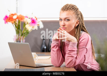 Nachdenkliche junge trendige blonde Frau mit geflochtenem Haar an einem Laptop in der Küche sitzt seitlich an der Kamera mit einem spekulativen Expres Stockfoto