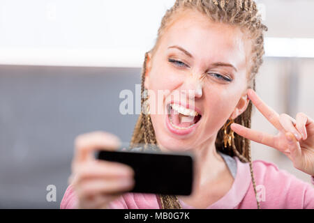 Junge attraktive blonde Frau, jubelten, als sie einen selfie auf ihrem Mobiltelefon, während Sie ein V-Zeichen mit Ihrem Finger in der Nähe zu 7/8-Ansicht Stockfoto