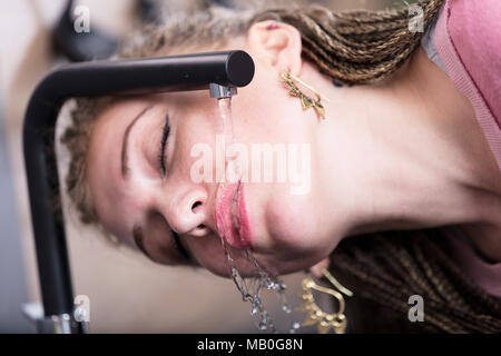 Attraktive Frau trinkt Wasser aus einem Hahn Holding ihr Gesicht unter dem flow Kaskadierung über Ihre geöffneten Lippen in das Becken oder Spüle in einer Nahaufnahme Kopf sh Stockfoto