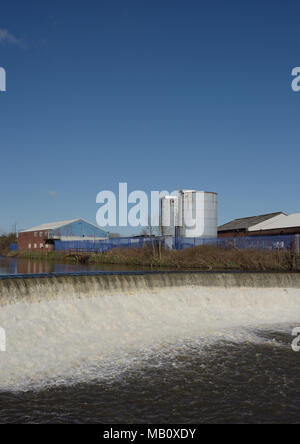 Wehr am Fluss Irwell verursacht eine turbulente Strömung von Wasser stromabwärts, zwei blauen Silos und Industriebauten Auf der Bank, radcliffe in Lancashire begraben. Stockfoto