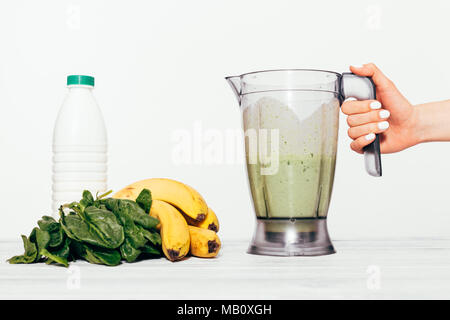 Woman's Hand hält frischen Smoothie in einen Mixer. Zutaten für Smoothie: Bananen, Spinat und Milch auf dem Tisch neben dem Prozessor auf einem weißen Stockfoto