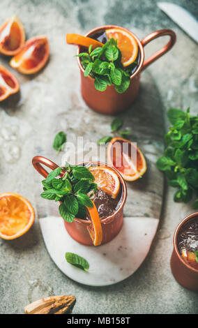 Blutorange Moscow Mule Alkohol Cocktails mit frischen Minzeblättchen und Eis in Kupfer Tassen an Bord im Zentrum über grauer Beton Hintergrund Stockfoto