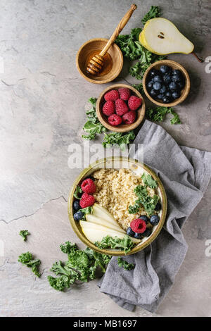 Vegan quinoa Porridge mit Grünkohl, Erdbeeren, Blaubeeren, in Scheiben geschnitten Birne, Honig auf Pendelarm in die Schüssel mit Zutaten oben und Stoff Serviette über graues Tex Stockfoto