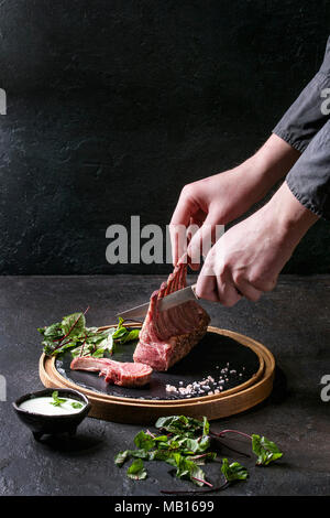 Des Menschen Hände Schneiden der Gegrillter Lammrücken mit Joghurt mint Sauce mit grünem Salat junge Rote Bete Blätter und rosa Salz auf runden hölzernen Schiefer serviert. Stockfoto