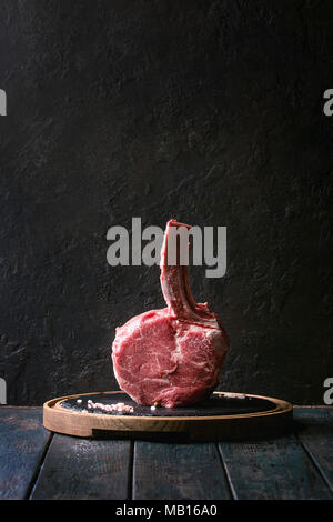 Roh ungekocht Black Angus Rind tomahawk Steak auf den Knochen mit Salz und Pfeffer auf runden hölzernen Schiefer Schneidbrett über dunkle Holzbrett serviert. Rus Stockfoto