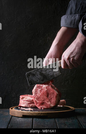 Des Menschen Hände schneiden Roh ungekocht Black Angus Rind tomahawk Steaks auf den Knochen vintage Metzger Cleaver auf runden Holz- Schiefer Schneidbrett über dunkel wird Stockfoto