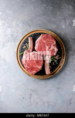 Roh ungekocht Black Angus Rind tomahawk Steaks auf Knochen mit Salz und Pfeffer auf runden Holz- Schiefer Schneidbrett über grau Textur Hintergrund serviert. T Stockfoto