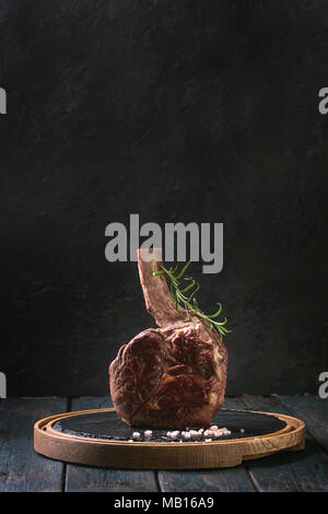 Gegrillte Black Angus Rind tomahawk Steak auf den Knochen mit Salz, Pfeffer und Rosmarin auf runde Schiefer Schneidbrett über dunkle Holzbrett Küche tabl serviert. Stockfoto