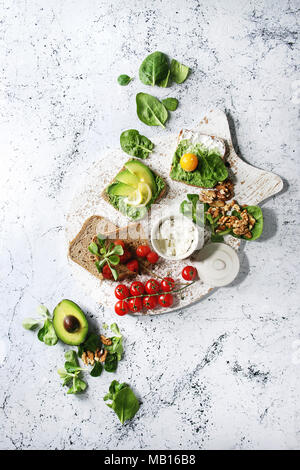 Vegetarische Sandwiches mit Avocado, Ricotta, Eigelb, Spinat, Tomaten auf Vollkorn Toastbrot auf weiß das Board mit Zutaten Abo Stockfoto