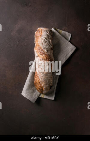 Laib des frisch gebackenen artisan Vollkorn ciabatta Brot auf Leinen über Dunkelbraun Textur Hintergrund. Ansicht von oben, kopieren. Stockfoto
