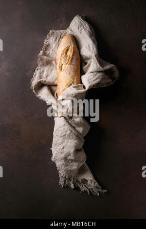 Laib geschnitten frisch gebackene artisan Baguette im Leinen über Dunkelbraun Textur Hintergrund gewickelt. Ansicht von oben, kopieren. Stockfoto