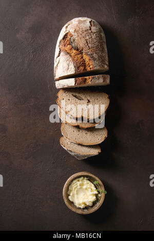 Laib des frisch gebackenen Artisan in Scheiben Roggenbrot mit Butter über Dunkelbraun Textur Hintergrund. Ansicht von oben, kopieren. Stockfoto