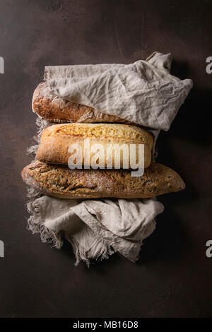 Vielfalt der Laibe frisch gebackene artisan Roggen-, Weiß- und Vollkornbrot auf Leinen über Dunkelbraun Textur Hintergrund. Ansicht von oben, kopieren. Stockfoto