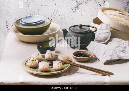 Asiatische Dampf potstickers Knödel von Garnelen, auf Keramik Teller serviert mit Sesam Soja Sauce, Essstäbchen, Teekanne, Tee gefüllt, Bambus Steamer über kitc Stockfoto