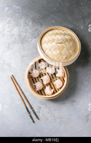 Asiatische Dampf potstickers Knödel gefüllt mit Garnelen in geöffnet Bambus Steamer mit Stäbchen über grau Textur Hintergrund. Ansicht von oben, Platz. Stockfoto