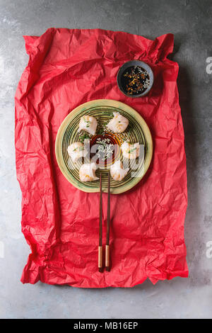 Asiatische Dampf potstickers Knödel gefüllt mit Garnelen, auf Keramikteller mit Sesam Soja Sauce und Essstäbchen auf zerknittertes Papier über Rot Grau tex serviert. Stockfoto