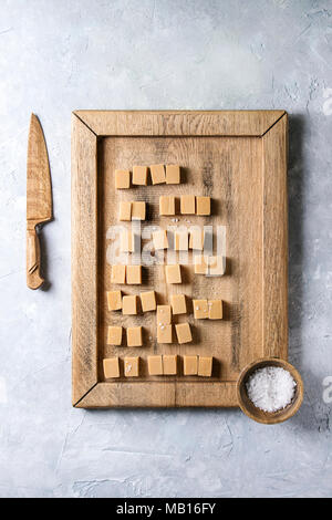 Gesalzene caramel fudge Candy auf Holz- Fach mit Fleur de in Holz Schüssel und Messer sel über grau Textur Hintergrund serviert. Ansicht von oben, Platz. Stockfoto