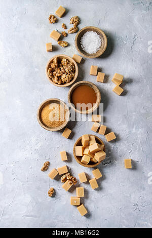 Gesalzene caramel fudge Bonbons mit Fleur de Sel, Karamell Sauce serviert, braunen Rohrzucker und karamellisierte Walnüsse in Holzschalen über grau Textur backgro Stockfoto