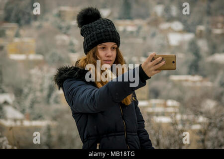 Recht jugendlich Mädchen im Schnee, nimmt selfie mit Telefon. Die Hälfte Körper Portrait von schönen kaukasischen Jugendmädchen, verschwommenes verschneite Stadt im Hintergrund. Stockfoto