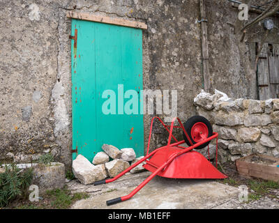 Rote Schubkarre auf dem Boden vor einer grünen Tür eines alten Hauses in Lubenice, Cres Kroatien liegen Stockfoto