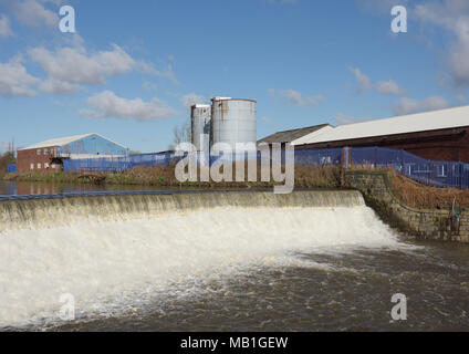 Wehr auf dem Fluss irwell verursacht turbulenten Wasserfluss stromabwärts, zwei blaue Silos und Industriegebäude am Ufer, radcliffe Bury lancashire uk Stockfoto