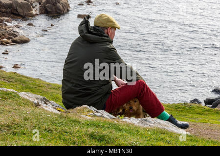 Ein Mann, gekleidet in einem flachen Kappe und Winterjacke sitzt auf der Spitze der Klippen mit einem kleinen Terrier Hund aus zu sehen. Denken gehen der Hund. Stockfoto