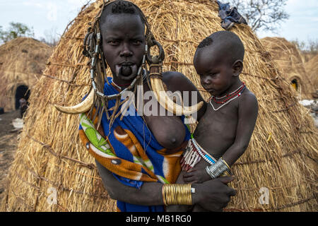 Omorate Aethiopien - Januar 24, 2018: Frau der Mursi Stamm mit traditionellen Schmuck und ihr Sohn mit der typischen Häuser der Mursi in Äthiopien. Stockfoto