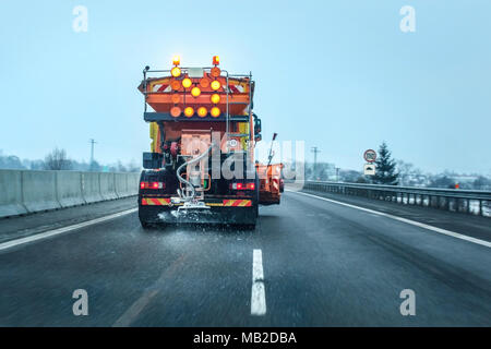 Blick aus dem Auto hinter Orange highway Maintenance truck Verbreitung Streusalz und Sand, kristalle fallen auf dem Eis bedeckten Asphalt. Stockfoto