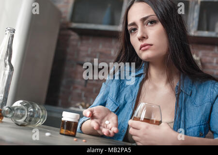 Junge weibliche Droge soziale Probleme am Tisch sitzen neben der Suche betonte unglücklich Holding Pille und Glas Whiskey süchtig Stockfoto