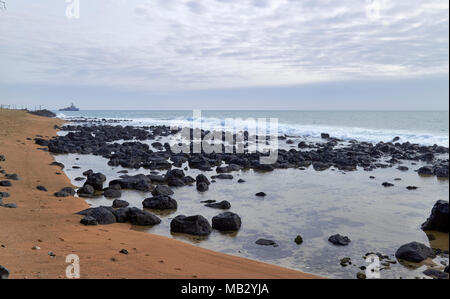 Den Strand entlang der alten Promenade von São Tomé Stadt spät zu einem warmen Nachmittag mit seiner orange Sand und dunklen vulkanischen Felsen in das klare Wasser. Stockfoto