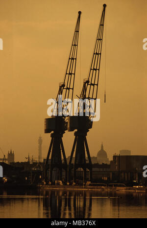 Isle of Dogs Docklands in London zeigen, Kräne und St Pauls Cathedral und der Post Tower in der Abenddämmerung. 1990 Stockfoto