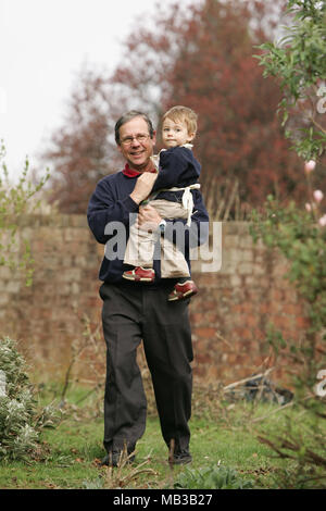 Ein alter Mann in seinem 50s lächelnd Pflege eine junge Alter 3-5 außerhalb in einem Garten. Die Beziehung zwischen den beiden ist Opa und Enkel Stockfoto