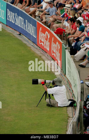 Ein professioneller Fotograf mit einem Teleobjektiv von Canon, montiert auf einem Stativ bei einem Kricketspiel sitzen auf dem in Feld vor der Werbetafel. Stockfoto