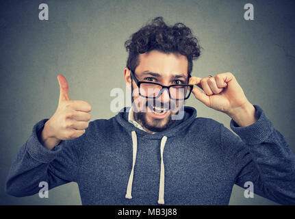 Jungen gutaussehenden Mann in Gläsern und Hoodie holding Daumen oben an der Kamera schaut. Stockfoto