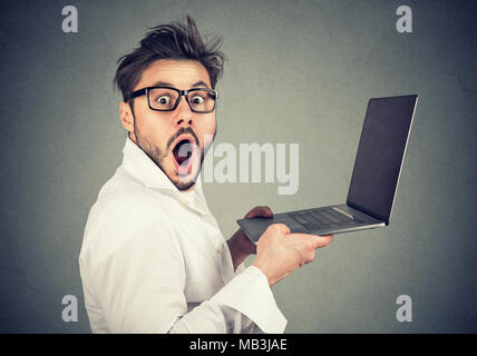 Seitenansicht des jungen Mannes in Brillen mit Mund auf Kamera geöffnet, während mit Laptop schockiert. Stockfoto