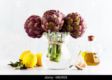 Frisches Bündel lila Artischocken in eine Karaffe mit Wasser auf weißem Hintergrund mit Zitrone, Olivenöl, Knoblauch. Frühling gesundes Essen Stockfoto