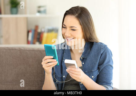 Glückliche Frau laden ein smart phone mit einem tragbaren Ladegerät auf eine Couch zu Hause sitzen Stockfoto