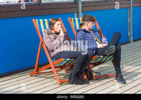Bournemouth, Dorset, Großbritannien. 7. April 2018. UK Wetter: Nach einem feuchten Start in den Tag die Sonne versucht, heraus zu kommen, obwohl es auf der kühlen Seite Besucher gehen nach Bournemouth Strand am Meer während der Osterferien zu genießen. Paar in Liegestühlen auf der Pier essen Eis Credit entspannen: Carolyn Jenkins/Alamy leben Nachrichten Stockfoto