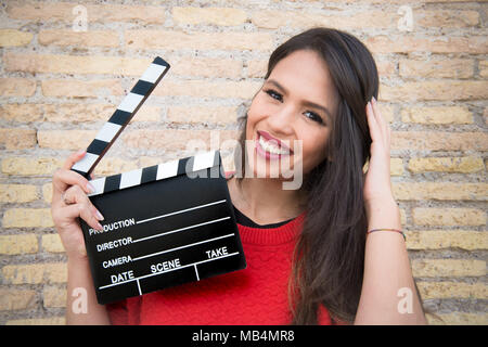 Porträt der jungen Hübschen Latina woman holding Filmklappe board mit Mauer im Hintergrund bei Kamera suchen Stockfoto