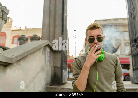 Junger stattlicher Mann mit grünen Kopfhörer und sunglasse genießen eine Zigarette auf der Straße. Stockfoto