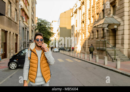 Junge Hipster mit orange Jacke, Sonnenbrille, Rucksack und sein Smartphone in der Stadt. Stockfoto