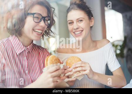 Zwei glückliche Mädchen mit Appetitlichen und Fett Cheeseburger Spaß und genießen die Zeit in Fast food Cafe Stockfoto