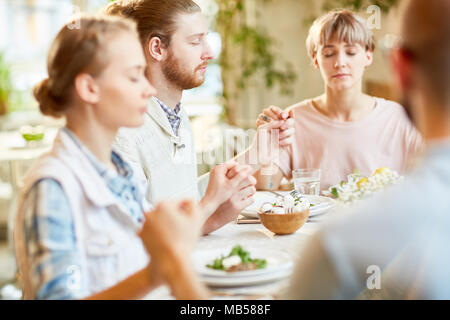 Gruppe von Freunden sitzen durch serviert Tabelle und halten die Hände beim Beten und Segen Essen vor dem Essen Stockfoto