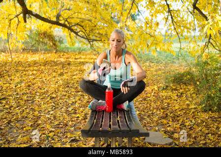 Attraktive Blondine Sportlerin erfrischend mit kalten Getränken können Sie über Ihr Smartphone. Auf der Bank sitzen in den öffentlichen Park. Gesunder Lebensstil Konzept. Stockfoto