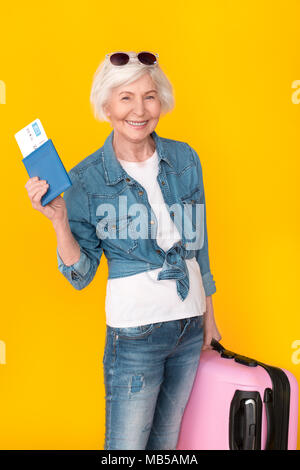 Im Alter von weiblichen Reisenden Studio auf gelben Hintergrund holding Pass und Flugticket isoliert tragen Gepäck Koffer suchen Kamera begeistert von neuen Trave Stockfoto
