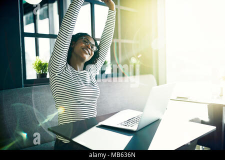 Junge afrikanische Frau lächelnd und strecken Sie die Arme, wenn sie alleine an einem Tisch online arbeiten mit einem Laptop Stockfoto