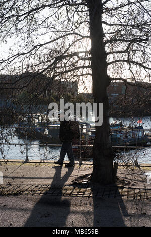 Silhouettenmensch spaziert entlang eines Flusspfades an einem Baum im Winterlicht vorbei, mit vertäuten Booten und Gebäuden im Hintergrund. Stockfoto