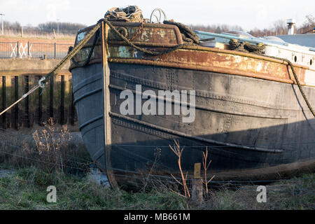 Ein rostgestreiftes Stahlboot liegt an Land, sein Rumpf wird mit dem Alter getragen, die Seile werden locker und halten den ruhigen Verfall und die Widerstandsfähigkeit des Lebens entlang der Gezeitenflüsse fest Stockfoto