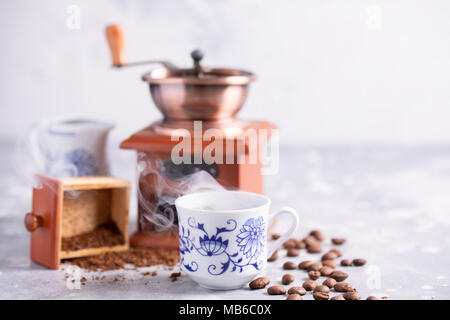 Körner von Kaffee fallen aus einer alten Kaffeemühle. Heißer Schwarzer Kaffee in einem schönen Porzellan Schale auf dem Tisch. Einen schönen Kaffee Komposition mit Stockfoto